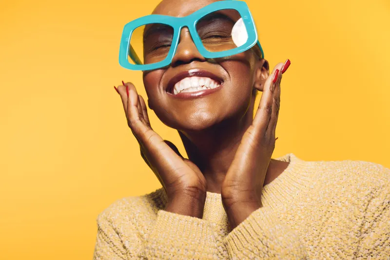 Smiling black woman with dark skin wearing large blue glasses and beige sweater poses joyfully on yellow background. Concept of happiness, fashion, and positive mood in studio.