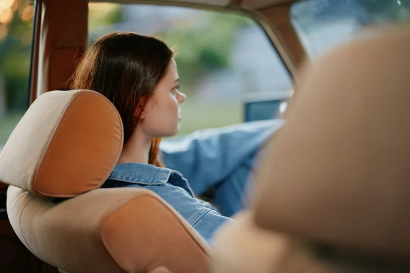 Young woman driving a vintage car, showcasing a relaxed and focused expression, dressed in a denim jacket, surrounded by a softly blurred outdoor background, warm colors.