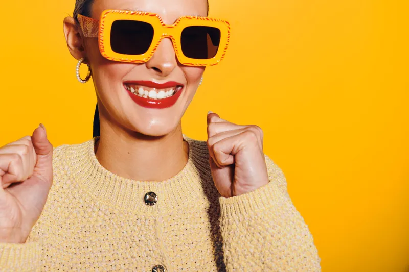 Happy woman with bright orange sunglasses and red lipstick smiles joyfully wearing a yellow sweater against a vibrant yellow background. Concept of fashion and positive mood.