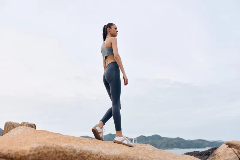 Fit Asian woman in activewear walking on rocky shore, showcasing healthy lifestyle and outdoor fitness, emphasizing nature and wellness