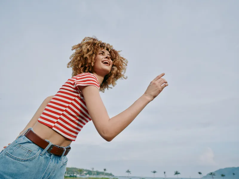 A joyful woman with curly hair runs at the beach under a blue sky, wearing a red striped shirt and denim jeans, capturing a vivid, carefree moment.