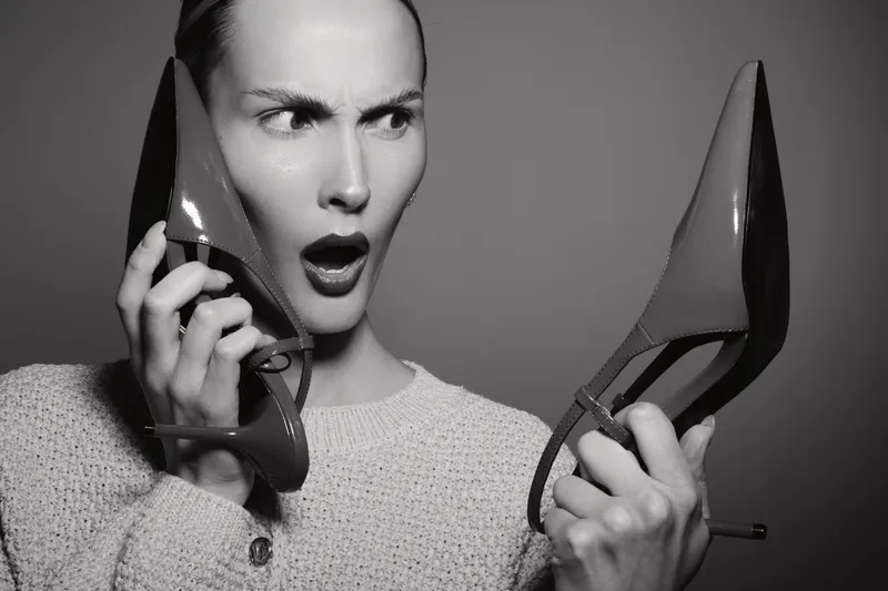 Surprised Caucasian woman with light skin holds red high heel shoes, looking shocked and confused. Monochrome portrait with strong facial expression and fashion concept.