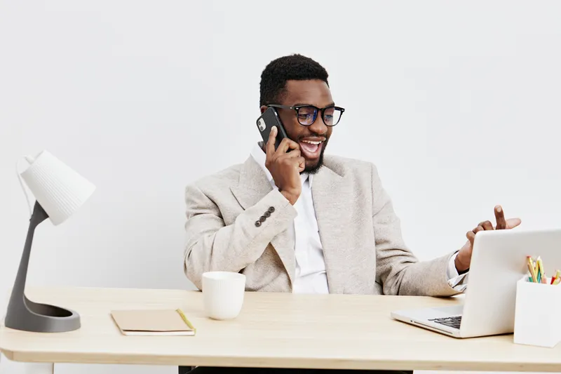 professional young African American man wearing glasses, beige blazer, using laptop and talking on mobile phone, smiling, work from home, modern office desk setup, corporate lifestyle concept