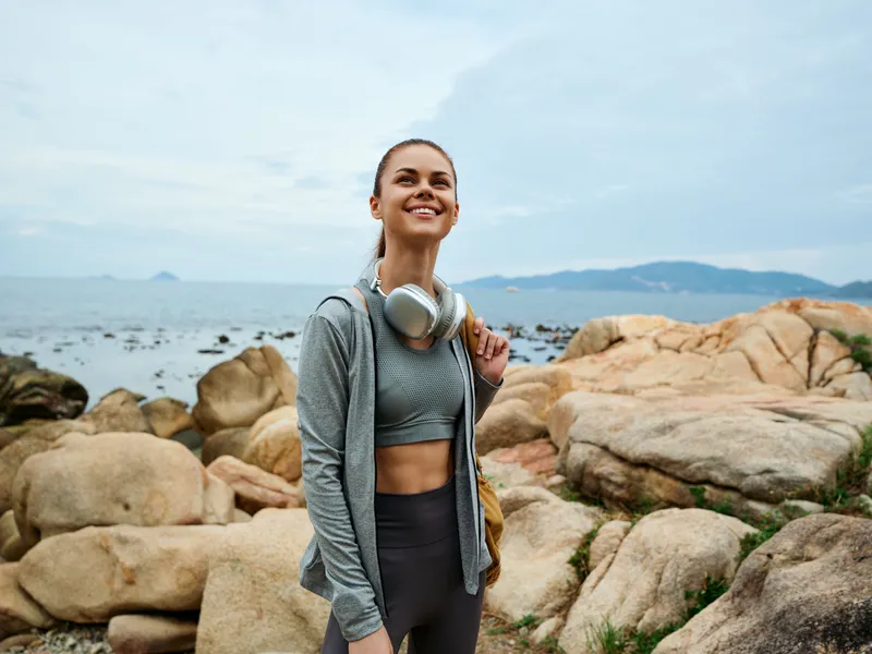 Young woman in activewear smiling on rocky beach with headphones, enjoying nature, ocean background, healthy lifestyle concept, fitness and happiness