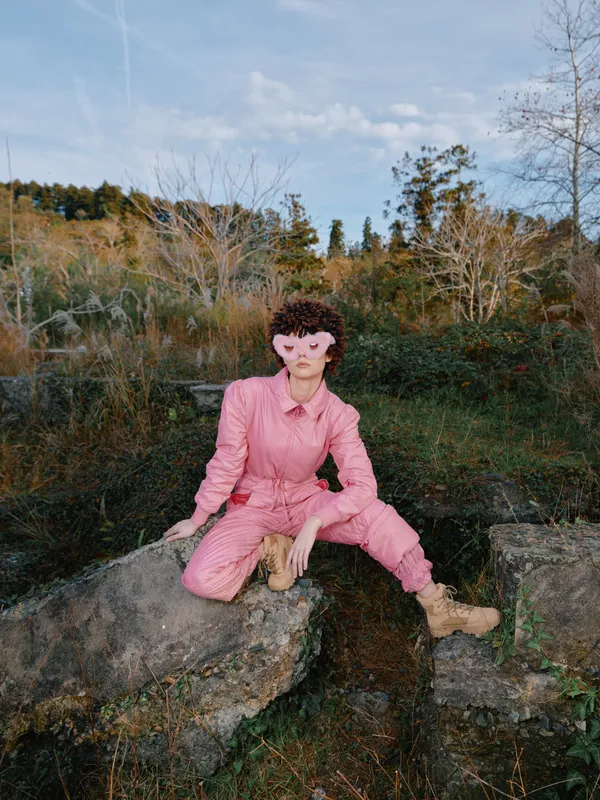 Outdoor person in a pink jumpsuit sits on rugged rocks amid trees and a clear sky, highlighting casual fashion, nature, and a relaxed pose.