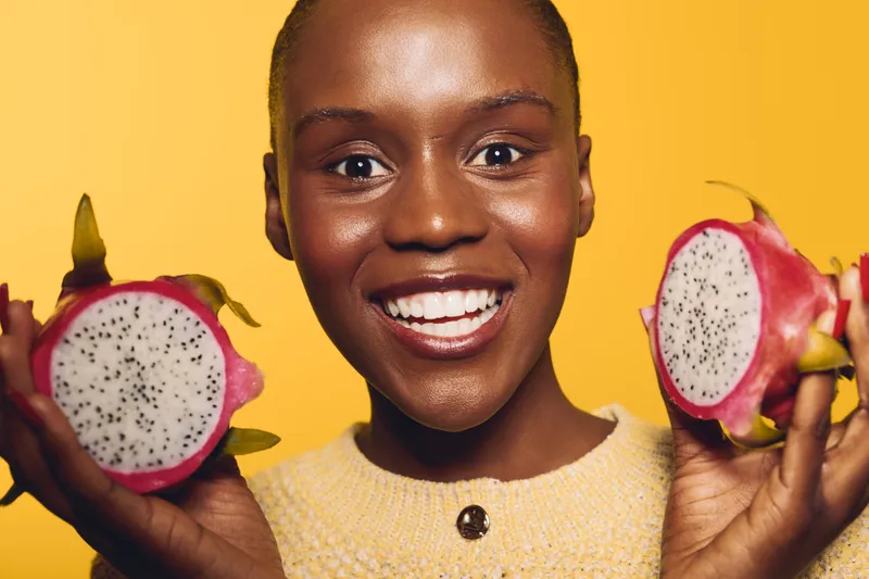 Smiling dark skinned African woman holds two halves of dragon fruit close to her face on yellow background. Concept of healthy lifestyle, fresh fruit, and natural beauty with vibrant colors.