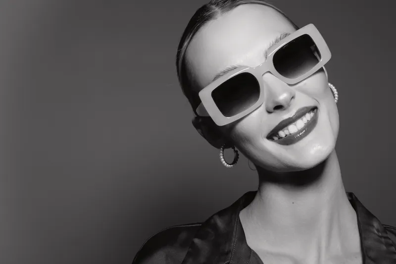 Smiling caucasian woman with pale skin wearing sunglasses and hoop earrings poses confidently against grey background in stylish black outfit.