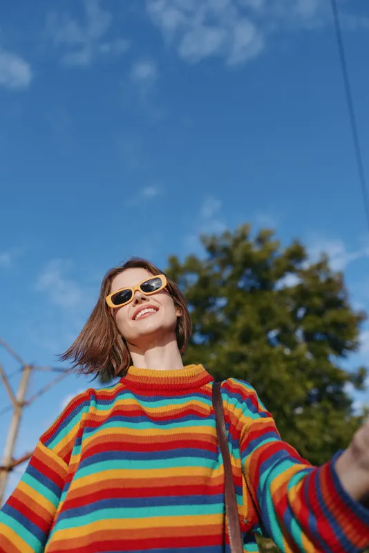 Woman in rainbow sweater wearing yellow sunglasses smiling outdoors, portrait with blue sky and sunlight. Happy young adult in colorful striped jumper, casual fashion and positive mood.