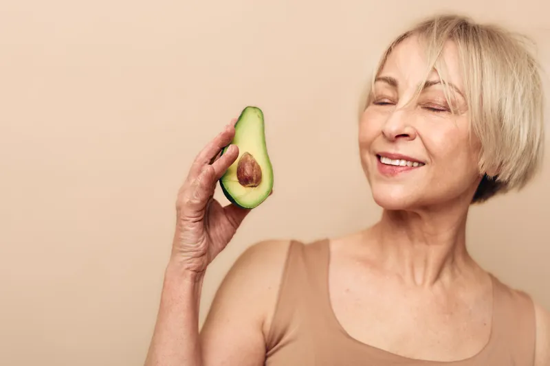 Smiling mature woman in beige tank top holds fresh avocado half near face with closed eyes. Concept of healthy lifestyle, natural nutrition, wellness, and organic food benefits for skin.