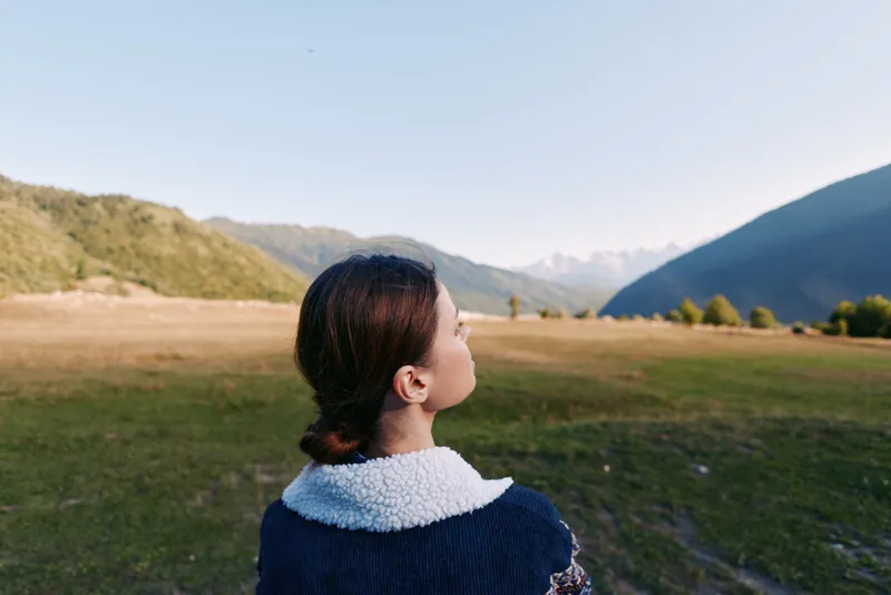 Woman with back to camera in mountain landscape and valley field, surrounded by nature and open outdoors, looking at distant mountains and a peaceful horizon.
