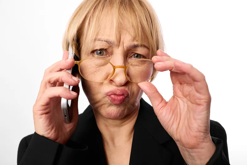 Elderly woman adjusts glasses while holding smartphone for phonecall, studio portrait on white background, communication, vision eyesight concept, isolated mature