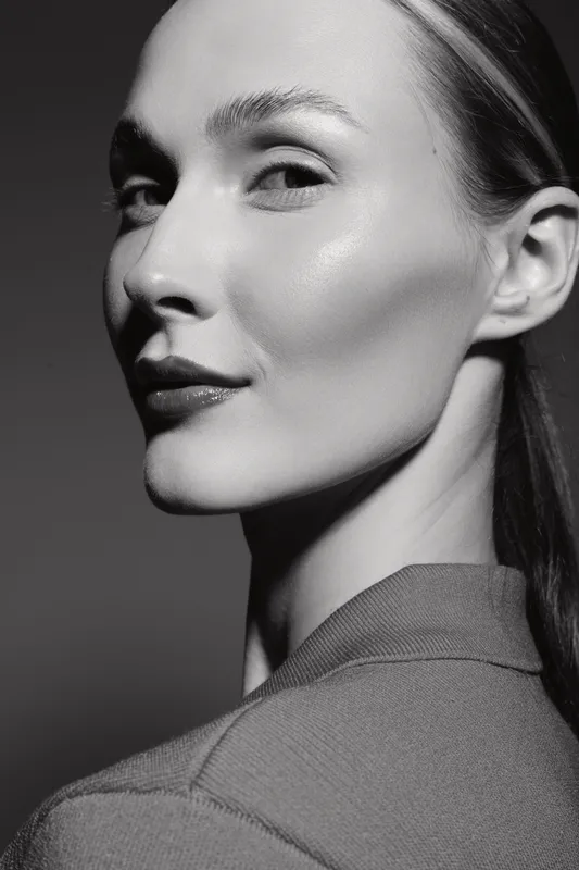 Close-up portrait of a young white woman with fair skin and long hair looking over shoulder. Studio lighting highlights facial features and smooth complexion.
