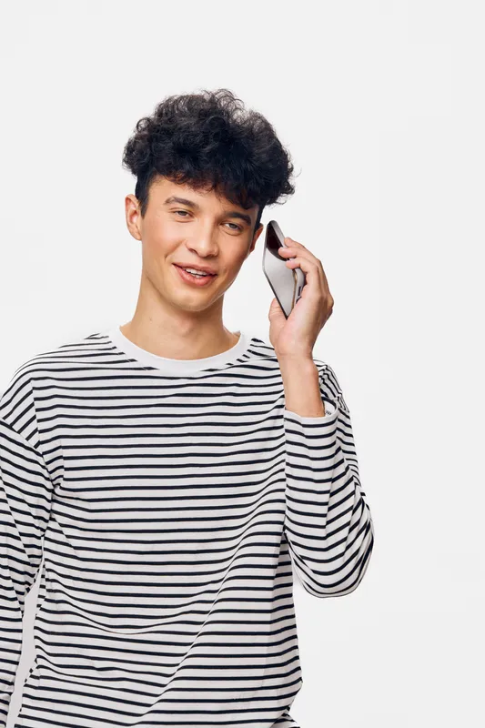 Young man with curly hair wearing striped shirt smiling while talking on smartphone, isolated on white background, casual lifestyle and communication concept.