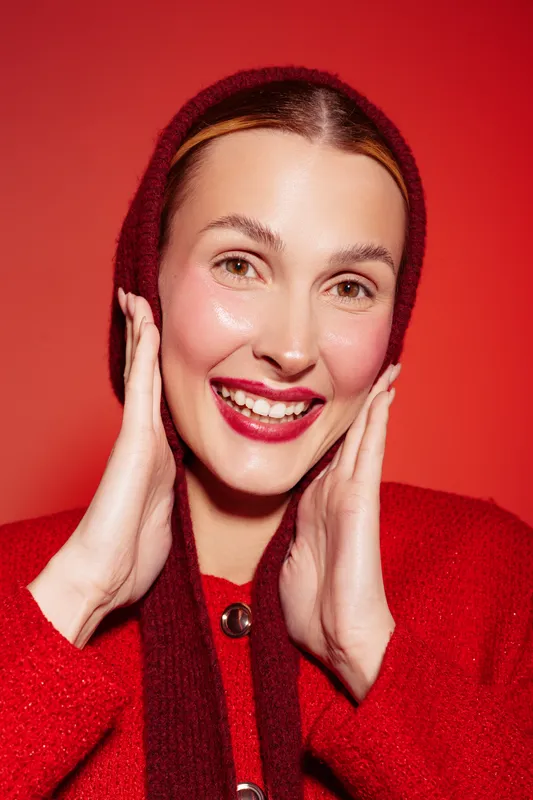 Smiling woman in red sweater and hood touches face with hands, showing natural makeup and healthy skin on red background. Concept of beauty, skincare, and happiness.