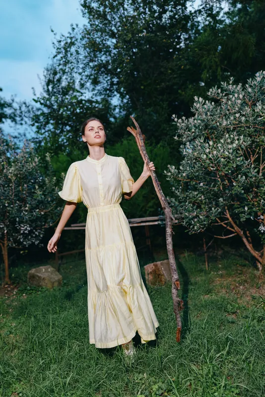 Woman in a pale yellow dress walks through a grassy garden holding a bare branch, sunlight filters through trees creating a serene outdoor scene