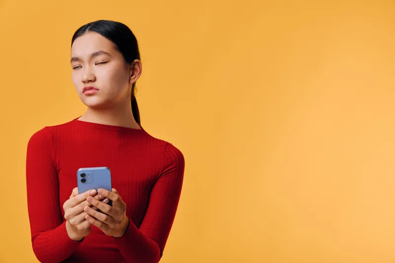 Young woman in red sweater holding smartphone with eyes closed against orange background. Female, casual style, digital device, communication and modern technology concept with thoughtful expression.