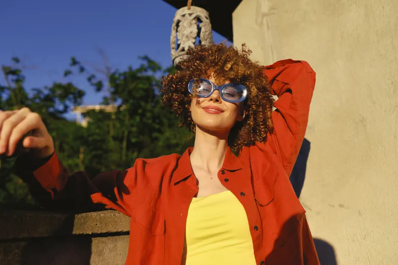 Bright woman lifestyle portrait outdoors in sunny weather, wearing a red jacket and yellow top, smiling confidently with sunglasses, casual fashion mood and relaxed pose