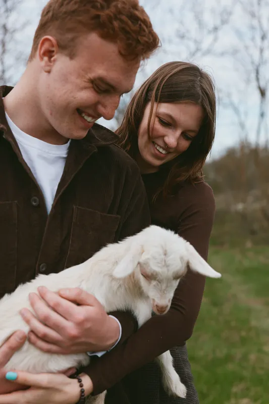 Joyful couple outdoors with a playful goat kid, smiling and embracing under soft light, natural colors, grassy backdrop, peaceful lifestyle moment, warm affection, candid scene