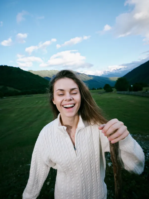 Smiling woman enjoying outdoor countryside with distant mountains, wearing a knit sweater, capturing a candid moment of joy and fresh air in natural light