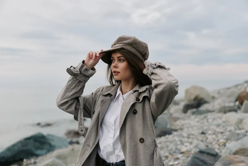 Young woman in stylish coat and hat stands on rocky beach under cloudy sky, looking to the side. Concept for fashion, autumn, and outdoor lifestyle photography.