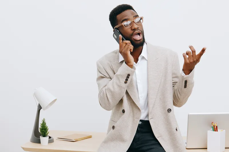 Professional young man with dark skin talking on mobile phone in office, expressing surprise or confusion, wearing beige blazer, white shirt, glasses, indoors against plain light background, business 