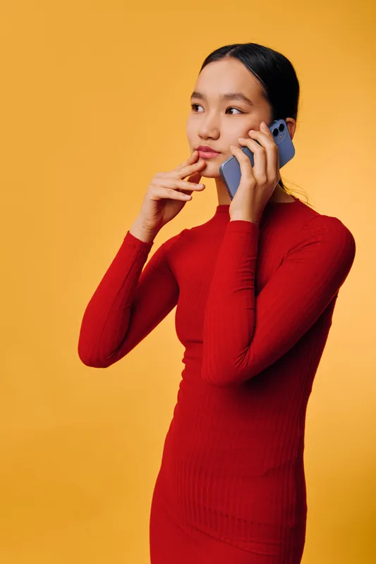 Young woman in red dress holding smartphone near ear and thinking with hand on chin, standing against yellow background in vertical portrait for communication and technology concept.
