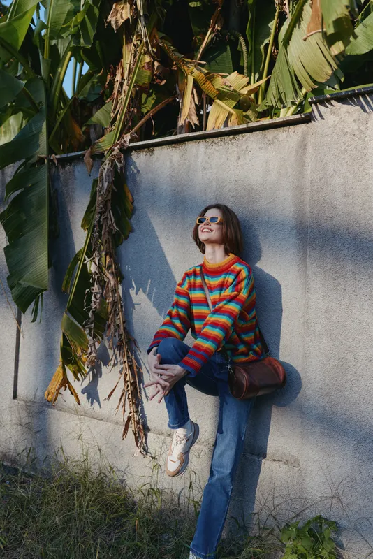 Young woman in a colorful striped sweater and sunglasses leaning against a concrete wall by banana leaves, smiling and wearing jeans with a brown crossbody bag for travel lifestyle.