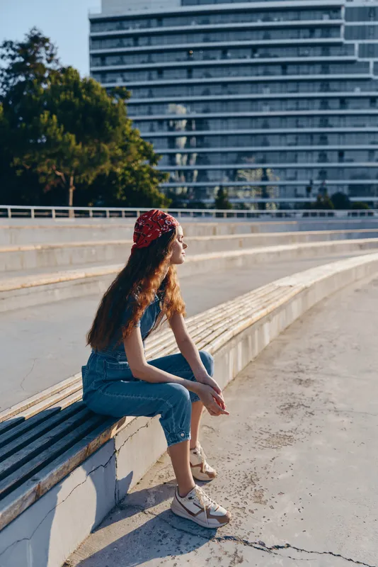 young woman sitting outdoors on wooden bench in casual jeans and red bandana enjoying sunny day with modern building and green tree background