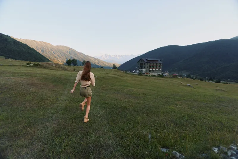 Active woman outdoors, running across a sunlit field toward distant mountains, with a rustic building in the background, depicting a carefree lifestyle