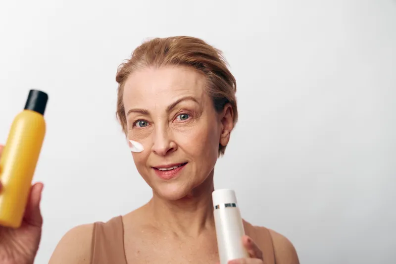 Mature woman with short hair applies white face cream, holding two cosmetic bottles. Skincare concept for aging skin, beauty routine, and self-care on light background.