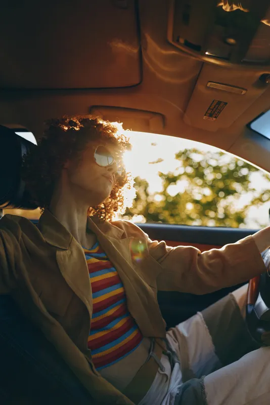 Woman driving a car with striped shirt and beige jacket. Sunlight fills the interior, creating a retro lighting mood for lifestyle and travel.