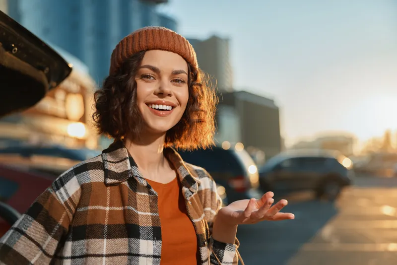 Woman smile in urban parking at golden hour, candid beanie portrait with sunlight backlight, plaid jacket and warm mood conveying authenticity, mindful living and emotional storytelling.