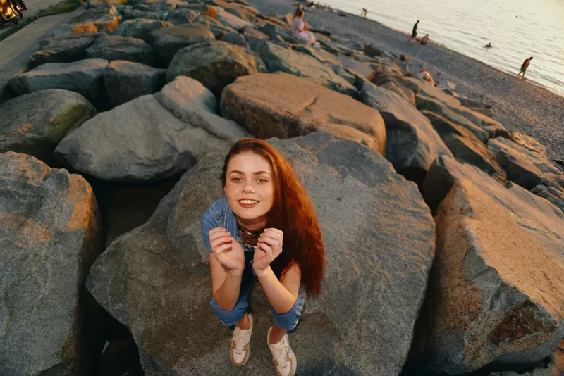 Lifestyle woman in modern boho-western denim street style smiling on rocky beach during warm golden hour with film color toning and casual relaxed mood
