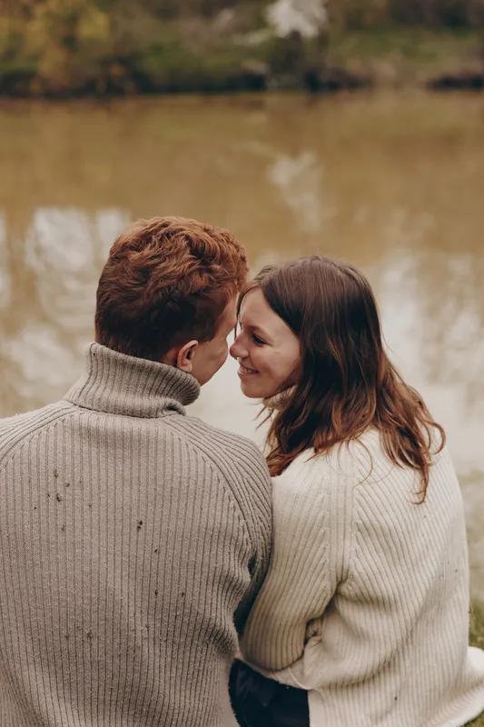 Two people wearing cozy sweaters share a quiet lakeside moment, smiling softly. Close embrace, warm tones, natural light. Concept of companionship, romance, and lifestyle in a serene outdoor setting.
