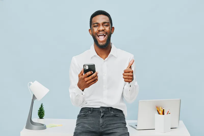 Smiling happy young man wearing white shirt holding mobile phone showing thumbs up in modern office workspace isolated on light blue background positive emotion concept