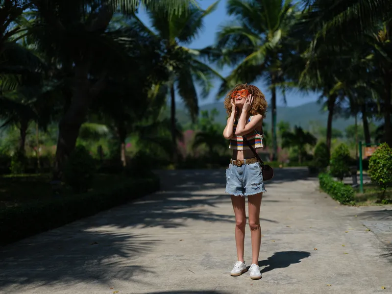 young woman with curly hair wearing sunglasses and denim shorts standing on sunny pathway surrounded by palm trees, enjoying warm tropical weather