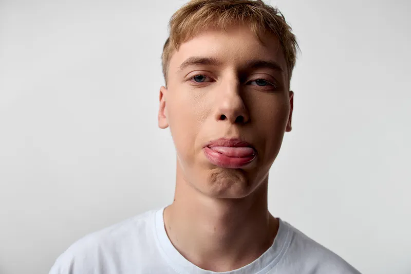 Young man with a playful expression sticks out his tongue in a casual shirt, closeup face against a light background, studio lighting, cheeky look and relaxed pose
