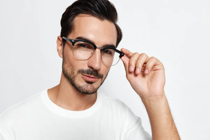Handsome young man wearing glasses adjusting his frames, looking confidently at the camera with a slight smile against a plain solid white background. Lifestyle concept.
