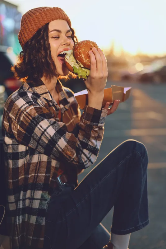 Woman eating burger outdoors at sunset, smiling in beanie and flannel. Candid lifestyle portrait with authenticity, golden hour glow, mindful living and emotional storytelling vibe.