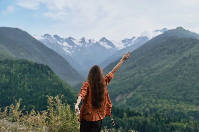 A person stands on a hillside with arms raised, gazing at a vast valley and snow kissed mountains, capturing freedom, adventure, and harmony with nature.