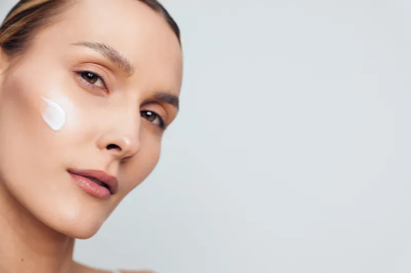 Close-up of a woman applying white cream to her cheek on a light background. Concept of skincare, wellness, and self-care. Soft lighting creates a calm, fresh mood.