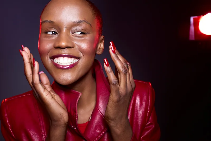 Smiling black woman with dark skin and short hair wears red leather jacket and red nail polish. Studio portrait with dark background and red lighting. Concept of fashion and happiness.