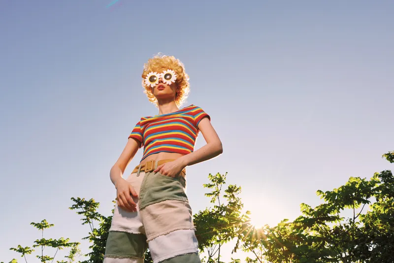 Cheerful woman outdoor wearing a striped shirt and floral sunglasses, standing confidently with sunlit outdoor backdrop and lush greenery, retro color mood, casual lifestyle moment in sunlight