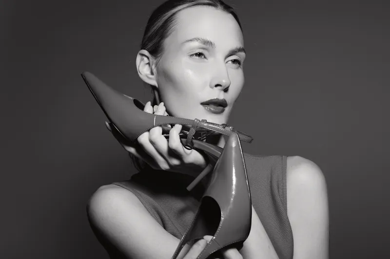 Elegant white woman with fair skin holds a pair of high heel shoes near her face in a monochrome studio portrait. Concept of fashion, style, and beauty accessories.