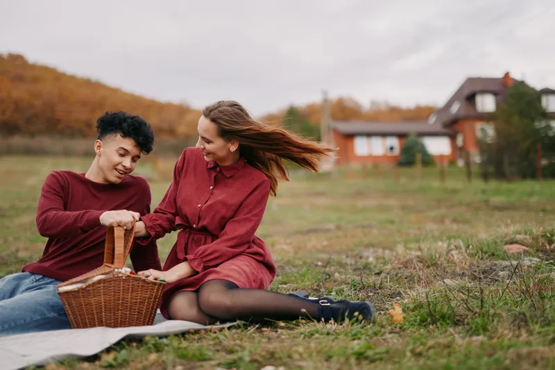 A young couple shares a genuine outdoor picnic on a cozy blanket, laughing as they unpack a wicker basket, creating a candid, believable moment of warm human connection.