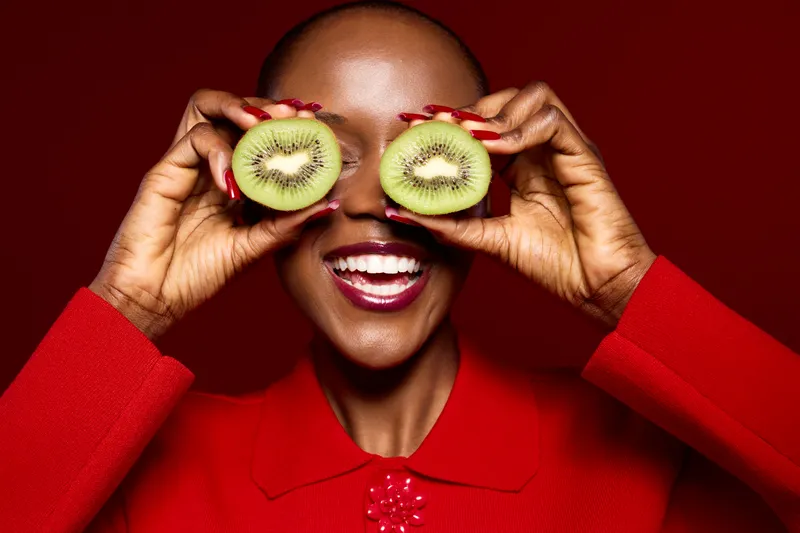 Smiling African woman with dark skin holds two halves of kiwi fruit over her eyes wearing red sweater. Bright studio lighting highlights joyful expression and fresh healthy concept with fruit.