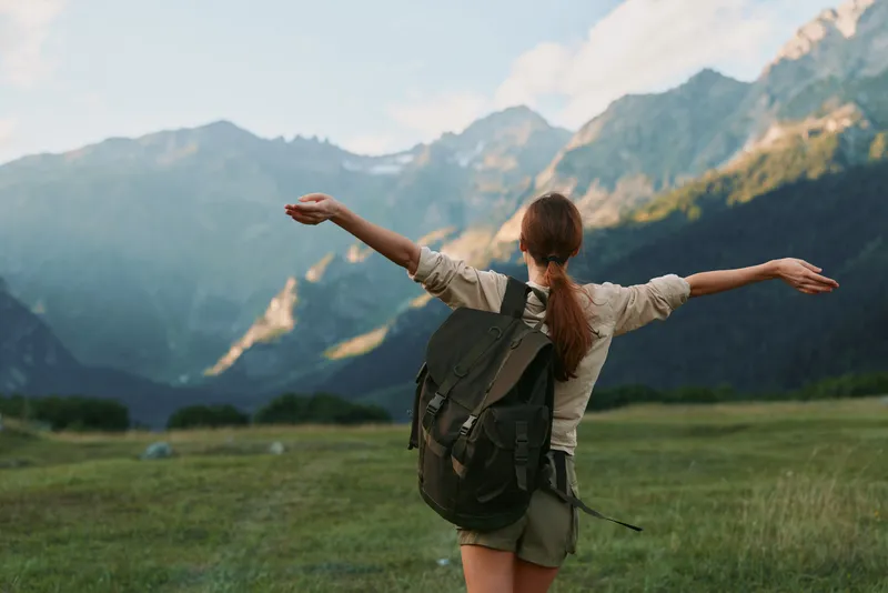 Young woman with backpack stands in a lush meadow with distant mountains, arms open, enjoying the fresh air and sense of freedom