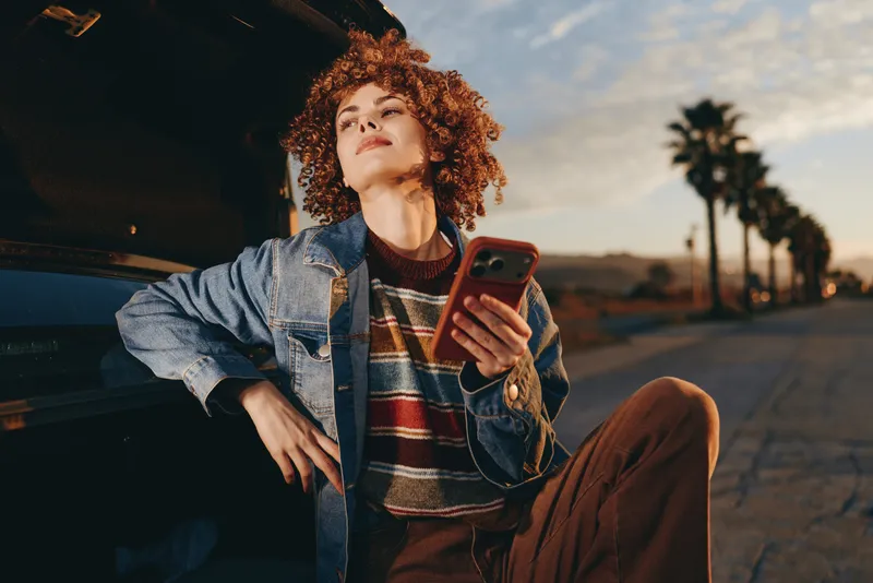 Woman lifestyle outdoor portrait, smartphone in hand, denim jacket, curly hair, leaning by car at sunset, casual relaxed mood on street road, warm evening sunlight, urban city dusk travel vibe