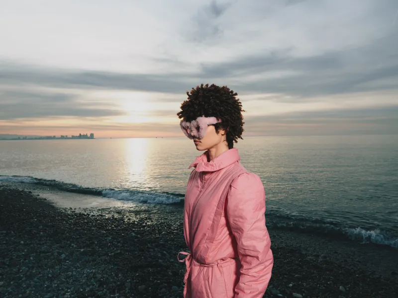 Person on a beach at sunset wearing a pink robe, calm sea in the background, poetic portrait with soft light, seaside breeze, reflective mood and tranquil scene
