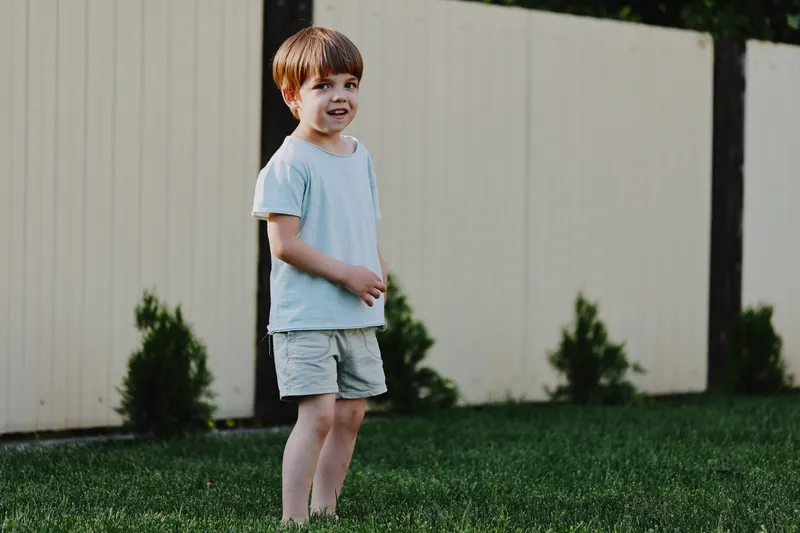 Happy boy standing on grass in a sunny backyard, wearing casual light blue clothing, enjoying a playful moment outdoors amidst green foliage.